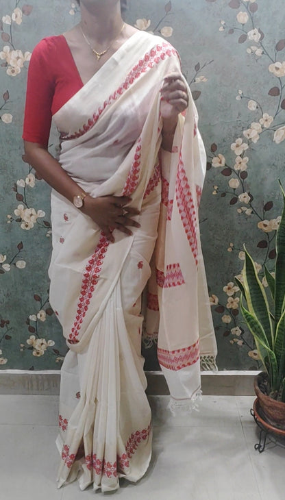 Woman wearing a white saree with red patterns in a room with floral wallpaper.