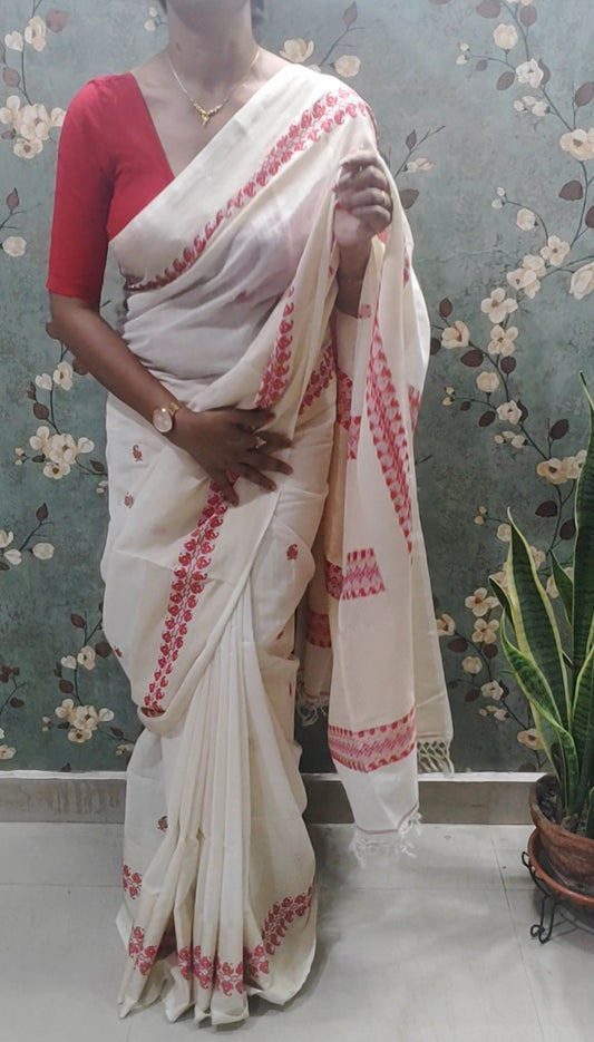 Woman wearing a white saree with red patterns in a room with floral wallpaper.